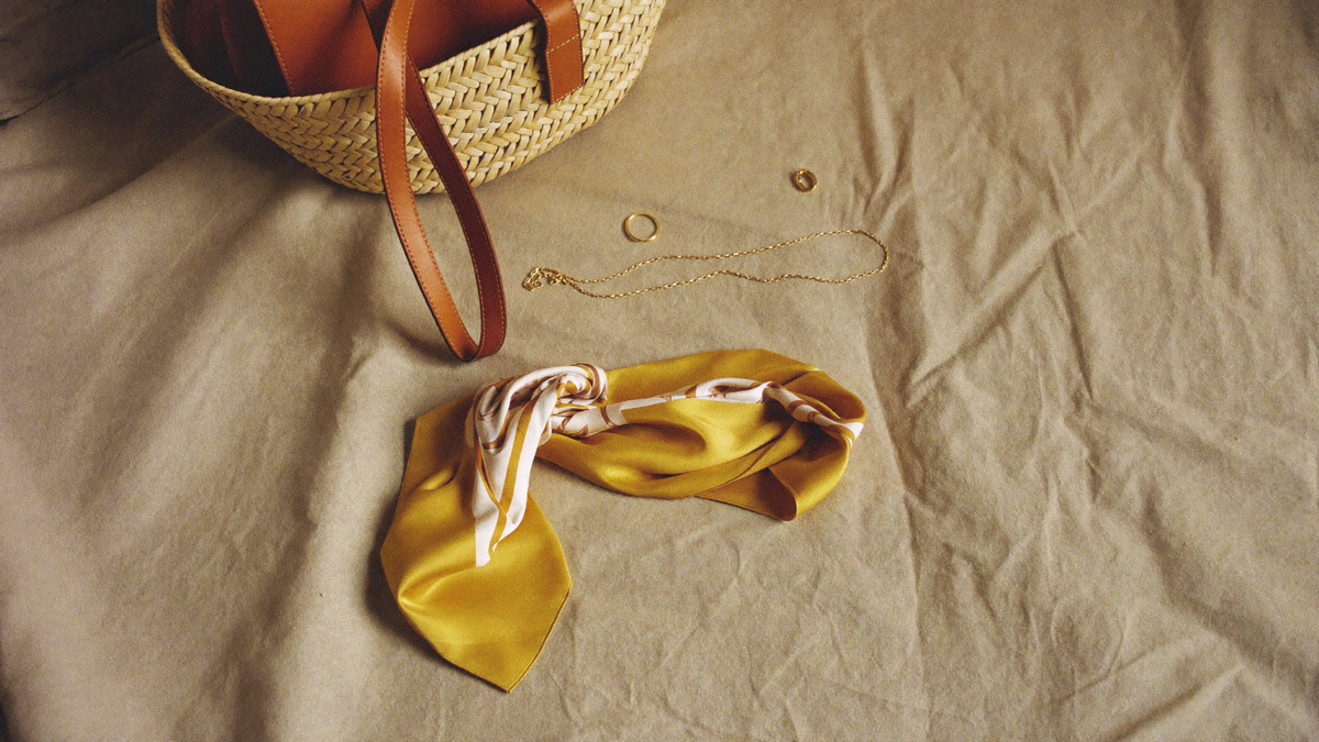 A woven straw and leather tote, fine gold chain necklace and folded silk scarf on natural linen cloth