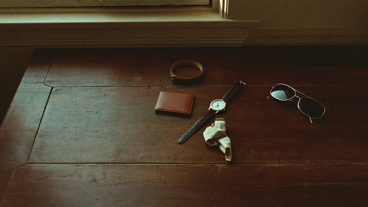 A leather bifold wallet, steel wristwatch, leather belt and metal-framed sunglasses on an aged oak table