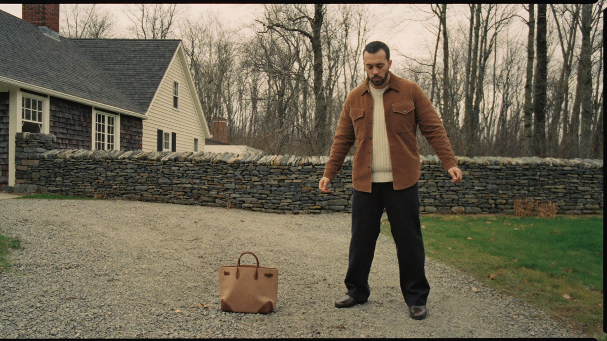 A man in a camel overshirt and cream knit on a gravel drive beside a shingled New England house in late autumn