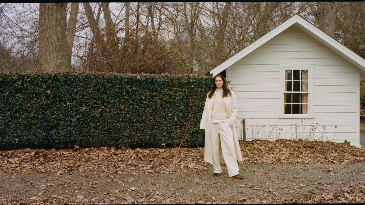 A woman in a long ivory wool coat on a leaf-strewn path beside a white clapboard New England house